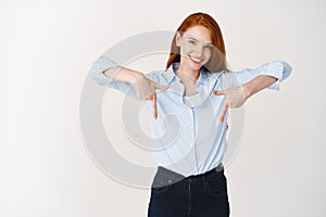 Business people. Young pretty redhead woman pointing fingers down, showing company logo and smiling, standing over white