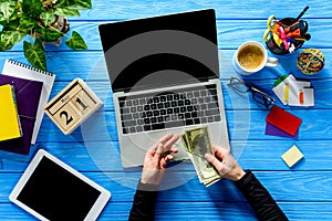 Business person tying dollars with rubber band by laptop on blue wooden table