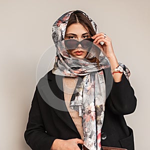 Business pretty elegant young woman in stylish silk scarf on head in fashionable sunglasses in black vintage coat posing outdoors