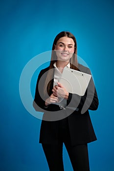 Business smiling woman in white shirt and black suit standing holding clipboard by her hands and looking forward, blue background