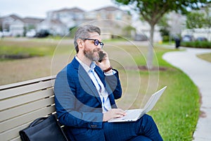 Business talk. Businessman talk on phone sitting on a bench in park. Businessman in suit call phone outside. Businessman