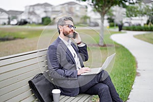 Business talk. Businessman talk on phone sitting on a bench in park. Businessman in suit call phone outside. Businessman