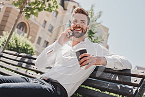 Business talk. Young man sitting in the park, talking on the phone and drinking coffee