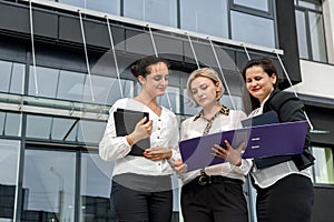 Business team. Three women in suits with folders and tablet posing outside building