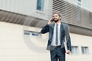 Business, technology and people concept - smiling businessman with smartphone talking over office building