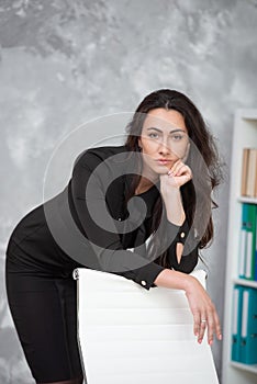 Business woman in a black dress is standing in an office, leaning on a chair