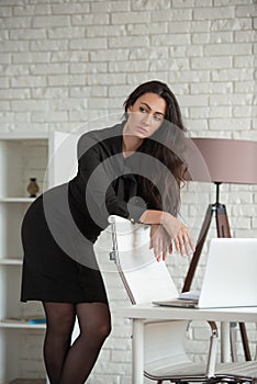Business woman in a black dress is standing in an office, leaning on a chair