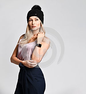 Business woman blonde in a satin blouse and dark trousers posing on a light background in a knitted hat with a clock on her hand.