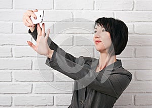 Business woman dressed in a gray suit taking selfie on smartphone or preparing to online broadcasting, stay in front of a white wa