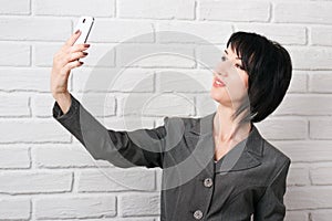 Business woman dressed in a gray suit taking selfie on smartphone or preparing to online broadcasting, stay in front of a white wa