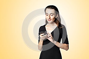 Business woman fatal woman in a black dress with red lipstick looks at her phone, isolated on a yellow background