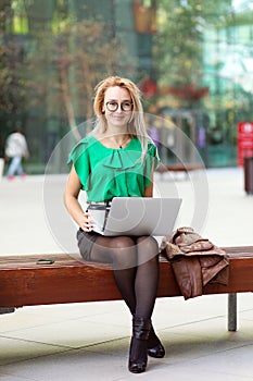 Business Woman with glasses sitting on bench outside street park business district drinking coffee and working on laptop