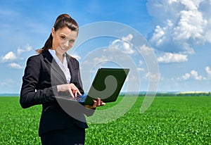 Business woman in green grass field outdoor work on laptop. Young girl dressed in black suit. Beautiful spring landscape with clou