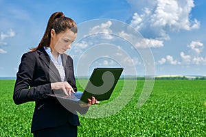 Business woman in green grass field outdoor work on laptop. Young girl dressed in black suit. Beautiful spring landscape with clou