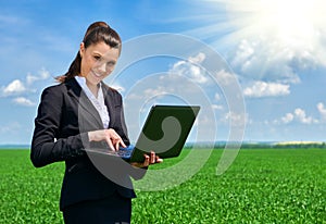 Business woman in green grass field outdoor work on laptop. Young girl dressed in black suit. Beautiful spring landscape with clou