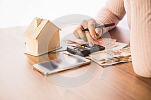 Woman hand counting money paper currency with smart phone,house model and calculator on desk, planing to buy or rent home