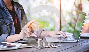 Business women hand holding light bulb on stack coins and working with computer on workplace. Creative ideas concept of saving mon