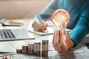 business woman hand holding lightbulb with coins stack on desk. concept saving energy