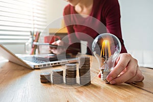 Business woman hand holding lightbulb with coins stack on desk. concept saving energy.