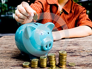 Business woman hand putting coin into blue piggy bank with gold coins pile growth graph, Saving money for future investment plan