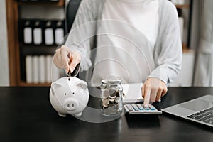 Business woman hand putting money coin into piggy bank with step of growing stack coins for saving money at modern office