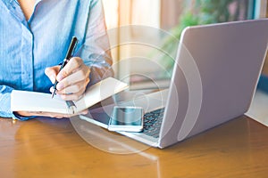 Business woman hand is writing on notepad with pen in office.On the wood table there are cell phones and laptops.