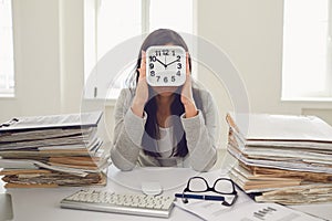 Business woman holding an alarm clock in front of her face while sitting at a table in the office