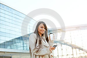 Business woman holding coffee and using  phone near office. Portrait of beautiful smiling female with phone, standing outdoors