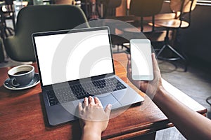 Business woman holding mobile phone with blank white screen while using laptop on wooden table in cafe