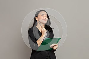 Business woman is imagining with pen on cheek while holding clipboard