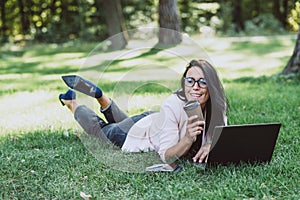 Business woman, lies in a summer grass park, using a laptop with a glass of coffee in hand. Remote work during quarantine. Woman