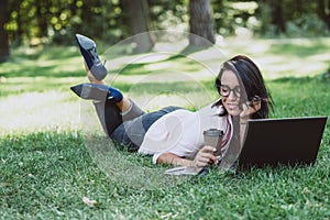 Business woman, lies in a summer grass park, using a laptop with a glass of coffee in hand. Remote work during quarantine. Woman