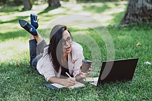 Business woman, lies in a summer grass park, using a laptop with a glass of coffee in hand. Remote work during quarantine. Woman