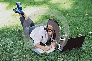 Business woman, lies in a summer grass park, using a laptop. Remote work during quarantine. Woman with coffee. Outdoors. Soft