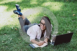 Business woman, lies in a summer grass park, using a laptop. Remote work during quarantine. Woman with coffee. Outdoors. Soft