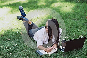 Business woman, lies in a summer grass park, using a laptop. Remote work during quarantine. Woman with coffee. Outdoors. Soft