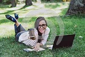 Business woman, lies in a summer grass park, using a laptop smiles and holds out a glass with coffee. Remote work during