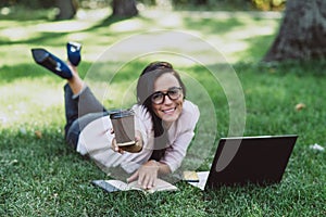 Business woman, lies in a summer grass park, using a laptop smiles and holds out a glass with coffee. Remote work during