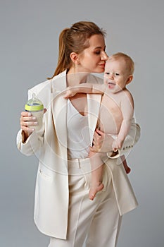 A business woman in a light suit holds a baby and a bottle of milk in her arms. A mother kisses a cheerful baby on a gray