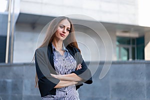 Business woman with long hair dressed black jacket standing outdoor near corporate office building hands folded