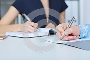 Business woman making notes at office workplace with colleague on the background. Man writing letter, list, plan, making