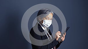 Business woman in protective mask with gray hair signs a document while holding tablet while standing next to dark wall