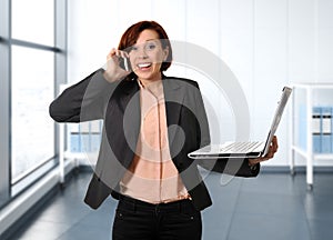 Business woman with red hair at work smiling with laptop computer talking busy on mobile phone