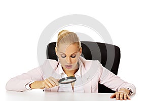 Business woman sitting behind the desk and looking into a magnifying glass