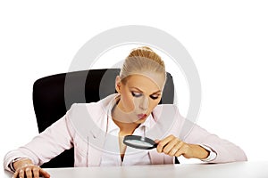 Business woman sitting behind the desk and looking into a magnifying glass