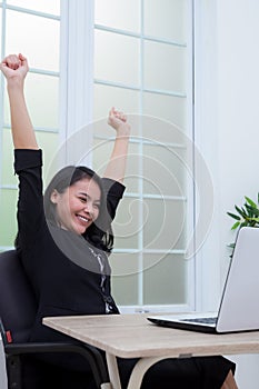 Business woman sitting on chair raising both hands with success expression in front of laptop