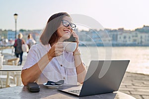 Business woman sitting in outdoor cafe with cup of coffee using laptop and smart phone