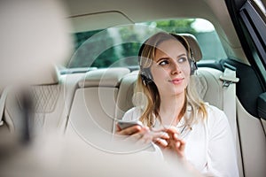 Business woman with smartphone and headphones sitting on back seats in taxi car.
