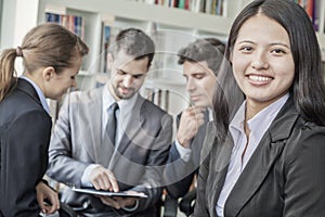 Business woman smiling and looking at the camera with her colleagues talking and looking down at a digital tablet in the