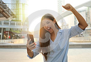 Business woman start the day with good news on her smart phone. Happy success woman celebrating outdoor cheering and raising her